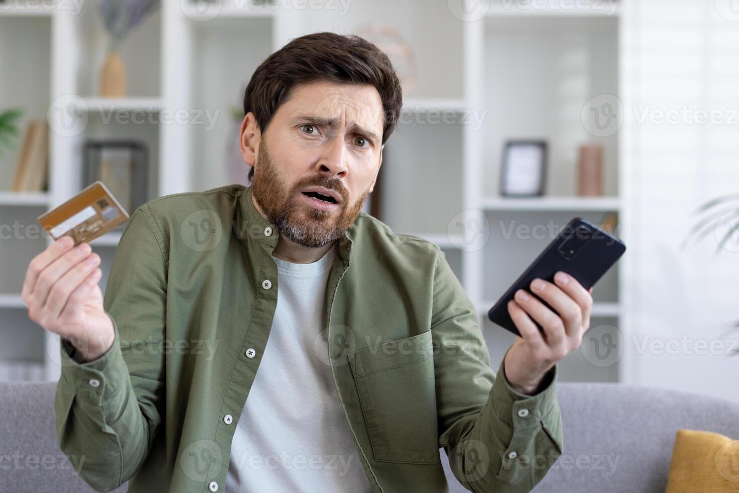 Confused adult man holding smartphone and bank card in hands while shrugging with worried expression. Uncertain customer having struggles with using ecommerce banking app for making online payments. photo