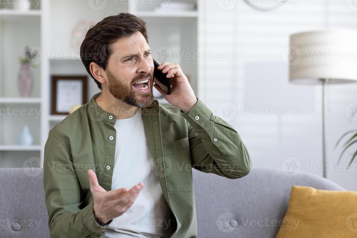 A bearded middle-aged man is engaged in an animated phone conversation, sitting alone on a sofa in a well-lit living room. photo