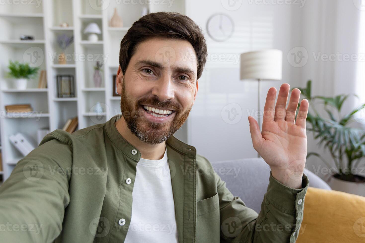 Adult male with stylish hairstyle waving hello with hand to camera while sitting on sofa over domestic interior background. Friendly guy in shirt greeting companion while starting new chat. photo