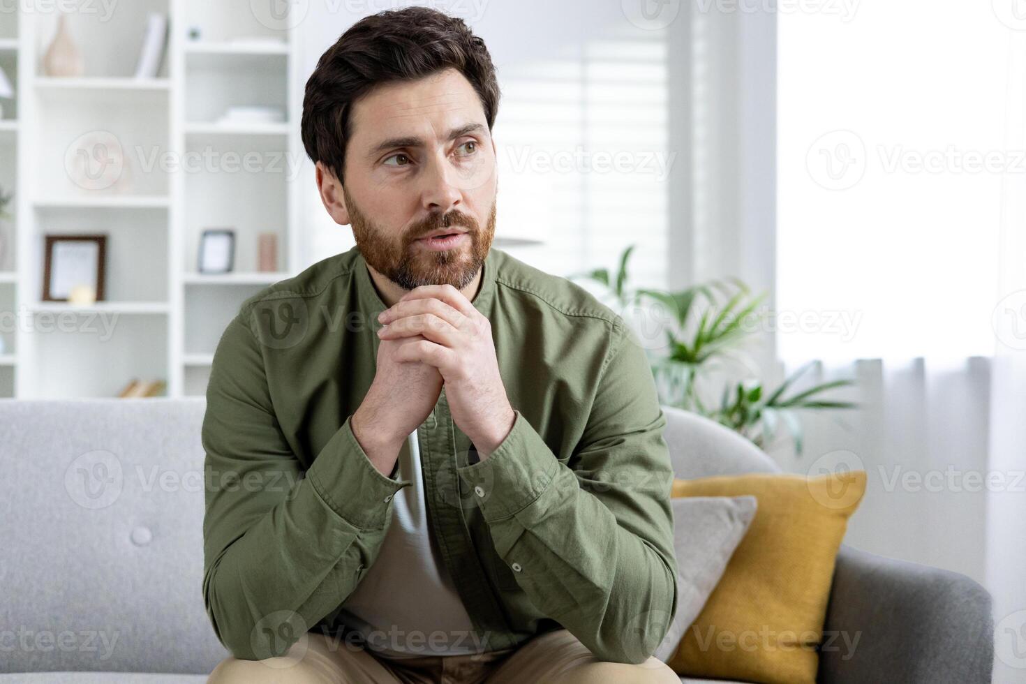Thoughtful man sitting on a sofa in a modern living room, deep in contemplation. Bright and airy home interior with bookshelf and plants. photo