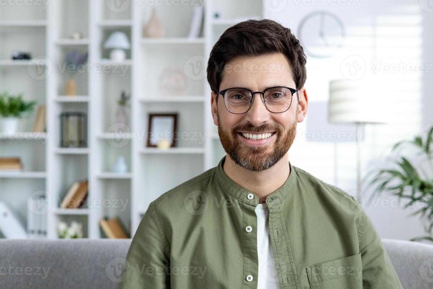Casually dressed man with glasses smiling at home, sitting on a couch with a well-organized bookshelf behind him photo