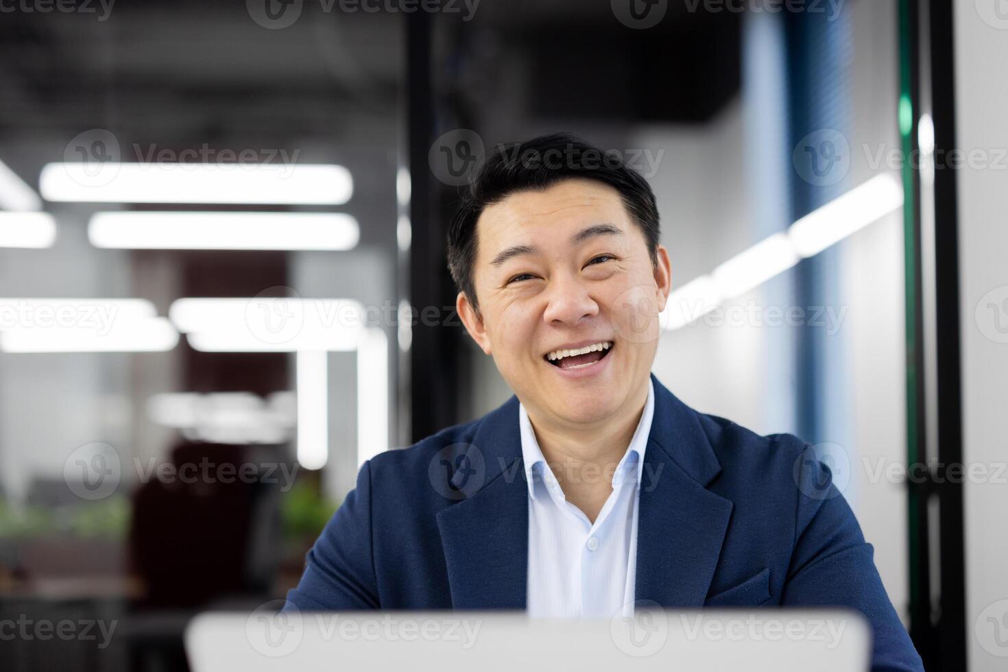 Portrait of happy japanese man in blue suit looking at camera with wide smile in office environment. Satisfied brunette employee doing data analysis of startup project and getting positive result. photo