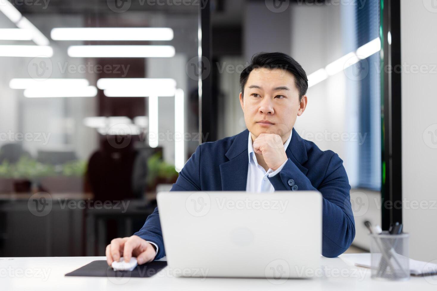 Pensive chinese male holding hand on chin while working on portable laptop with remote control mouse in spacious cabinet. Thoughtful man dressed in business suit having idea at personal workplace. photo