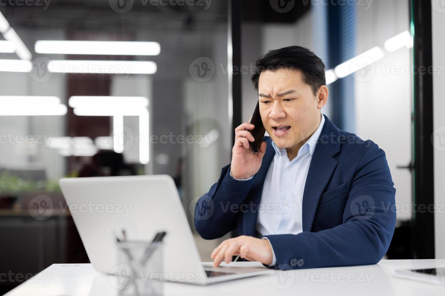 Angry grumpy boss shouting while talking on the phone, experienced asian man in business suit with phone in hands working inside office with laptop, annoyed businessman. photo