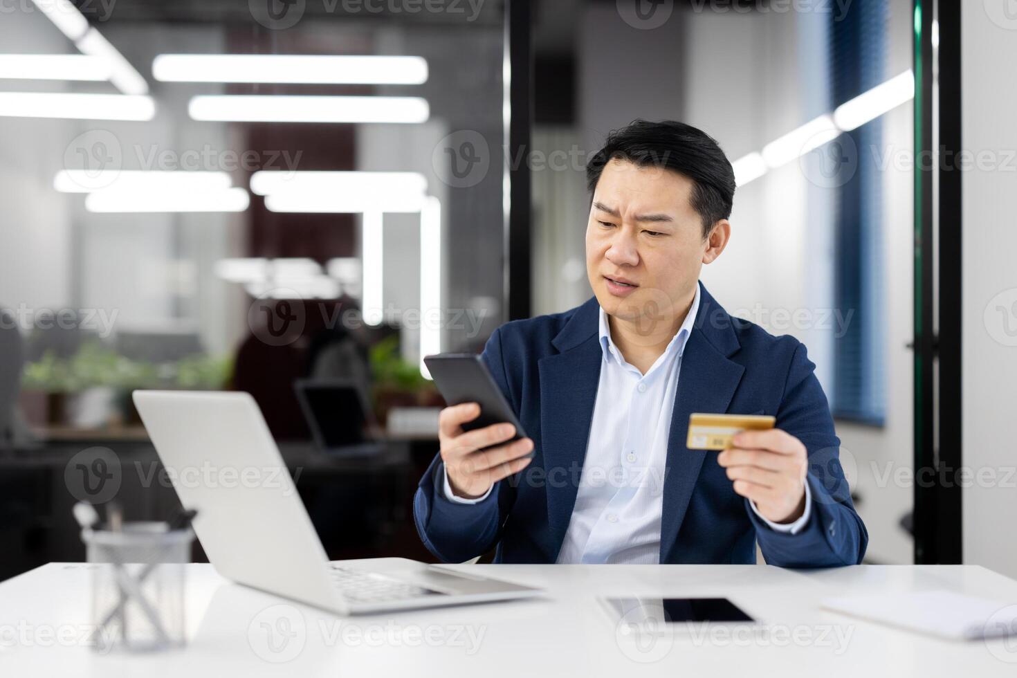 Perplexed Asian businessman in office looking at smartphone while holding a credit card, depicts online payment issues or security concerns. photo