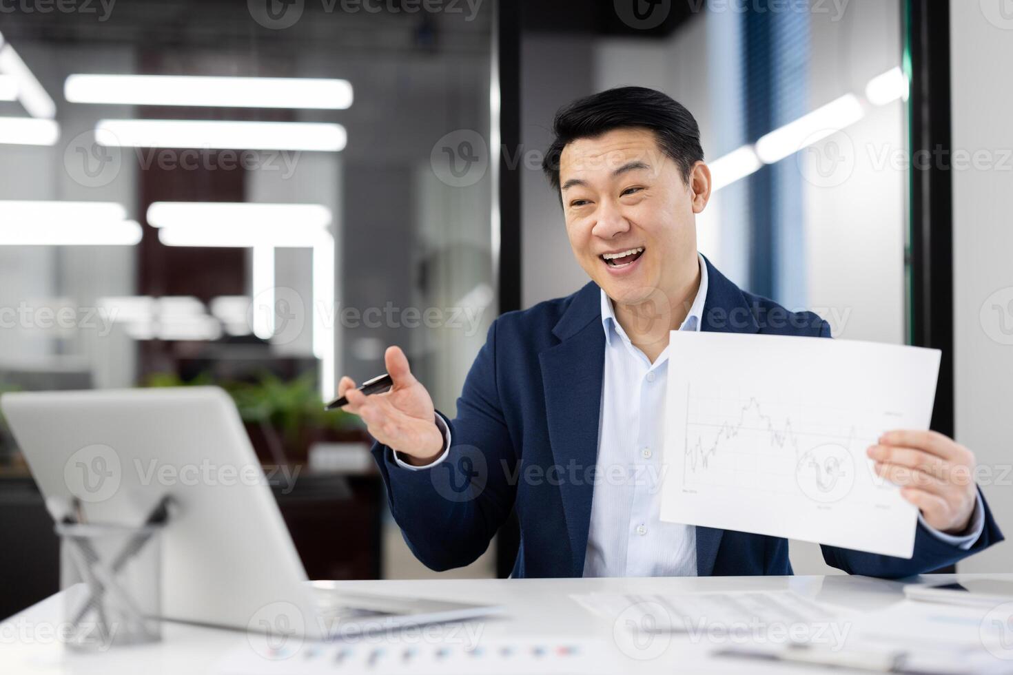 Confident Asian businessman in a blue suit smiling and holding a financial growth chart during a presentation in a modern office. photo