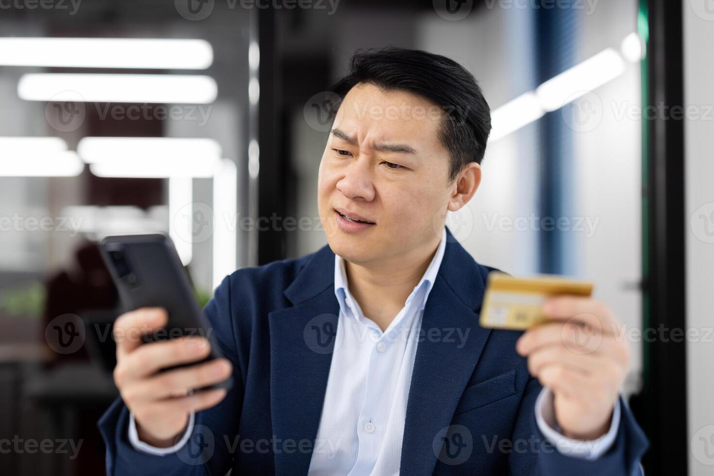A middle-aged Asian businessman looks confused and concerned while comparing his credit card to his smartphone in an office setting. Concept of financial fraud or banking error. photo