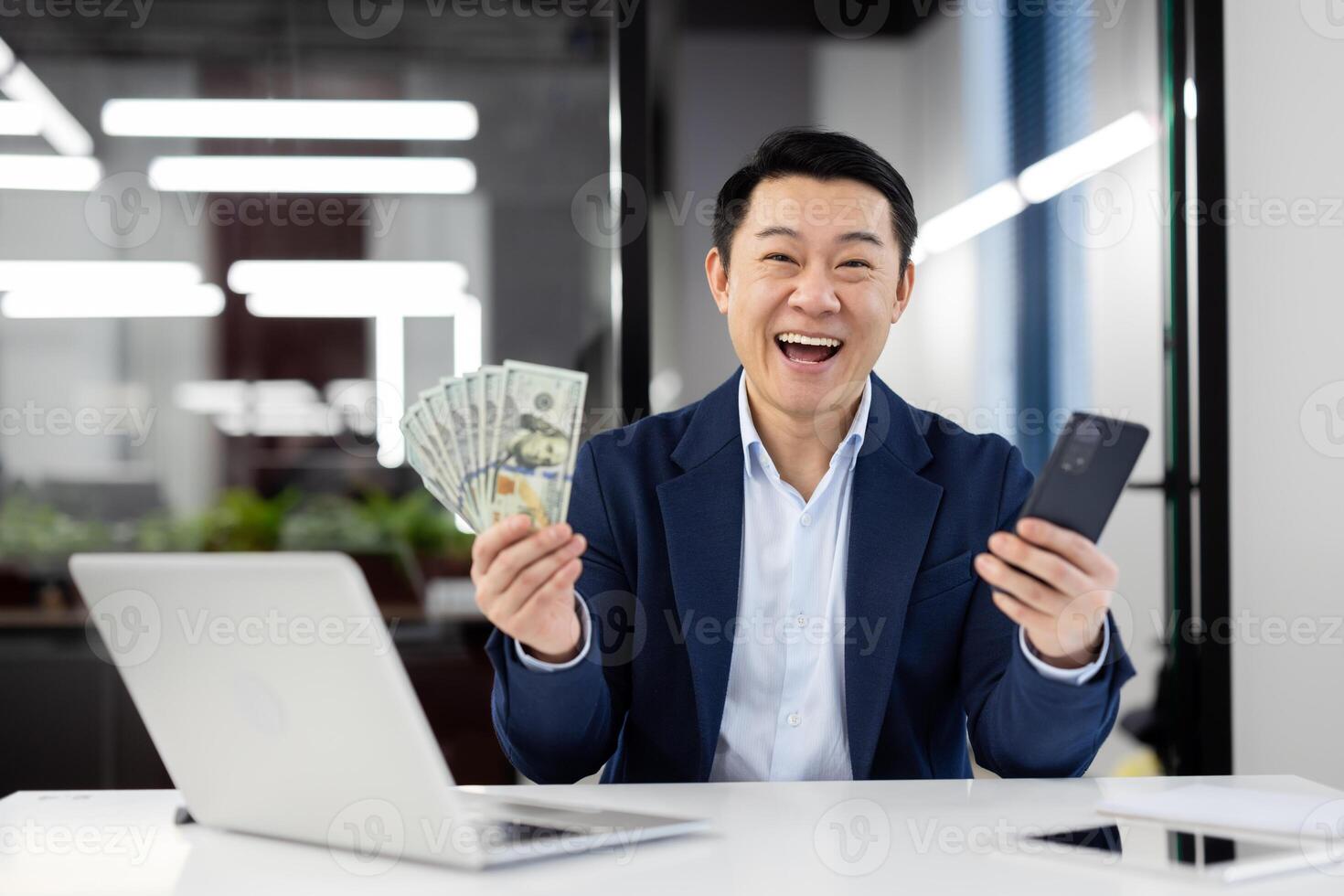 Joyful businessman in suit at office desk laughing with money in hand and a smartphone, expressing success and wealth. photo