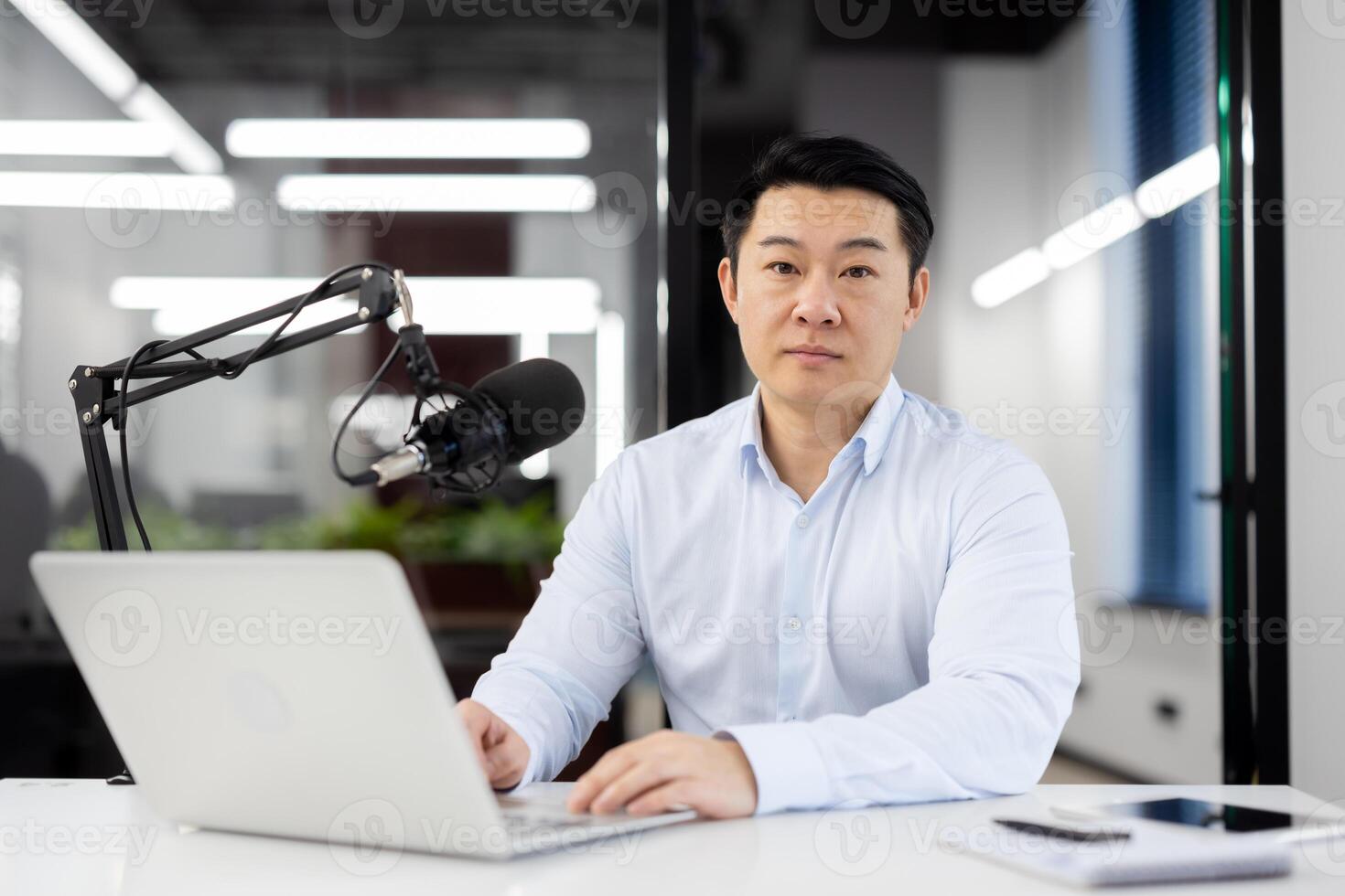 Portrait of a serious young Asian man in a white shirt sitting in the office at a desk, working with concentration on a laptop, looking confidently at the camera. photo