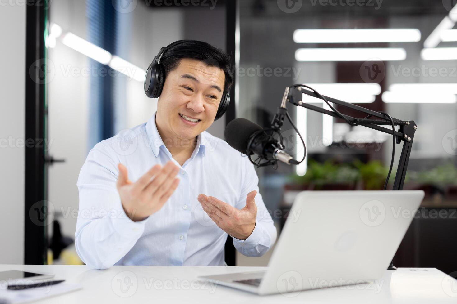 Smiling young Asian man wearing headphones sitting in office in front of desk with microphone and talking on call on laptop gesturing with hands. photo