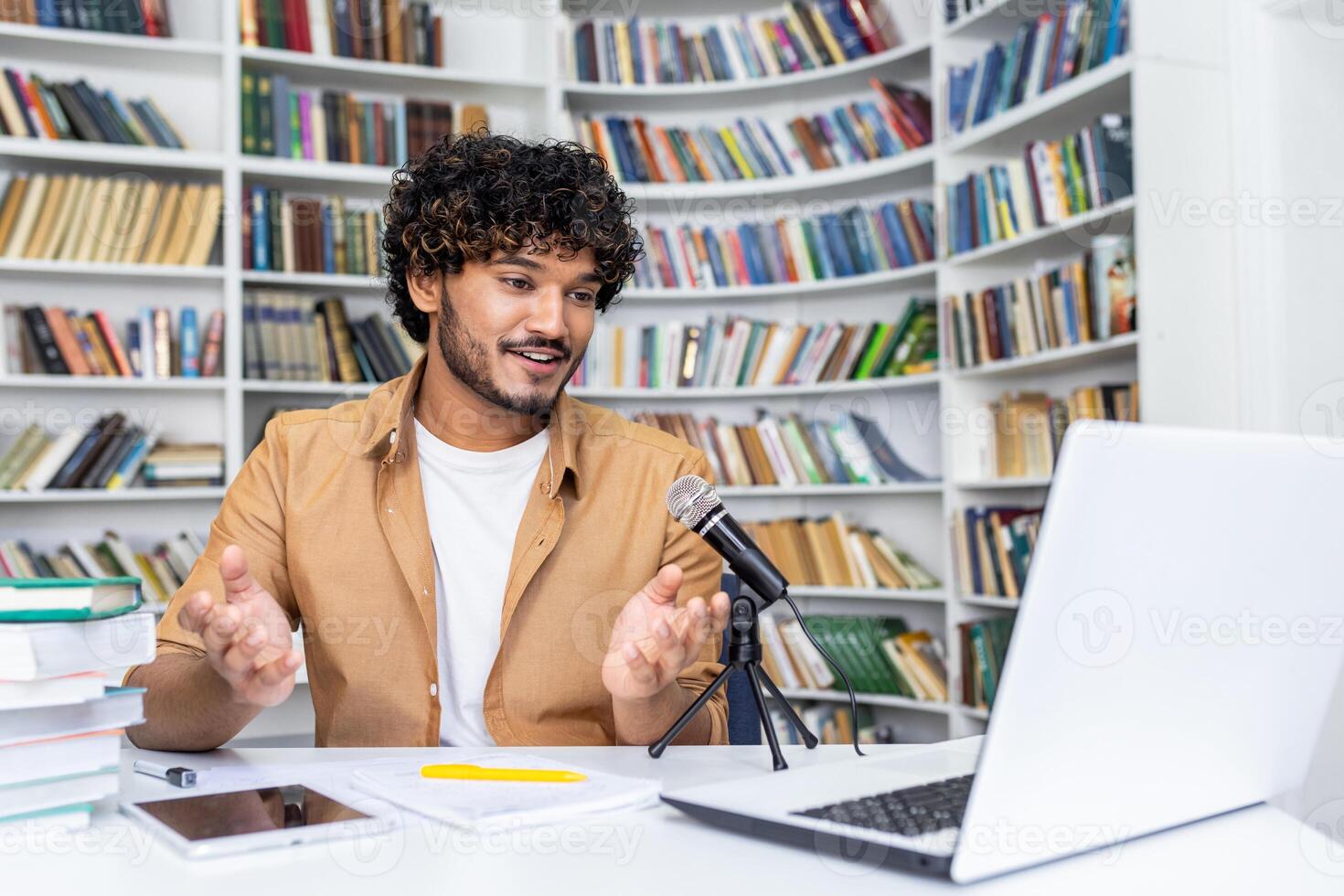 A cheerful young adult with curly hair talks into a microphone in a home office setup, engaging in a lively webinar or podcast, with a laptop and bookshelves in the background. photo