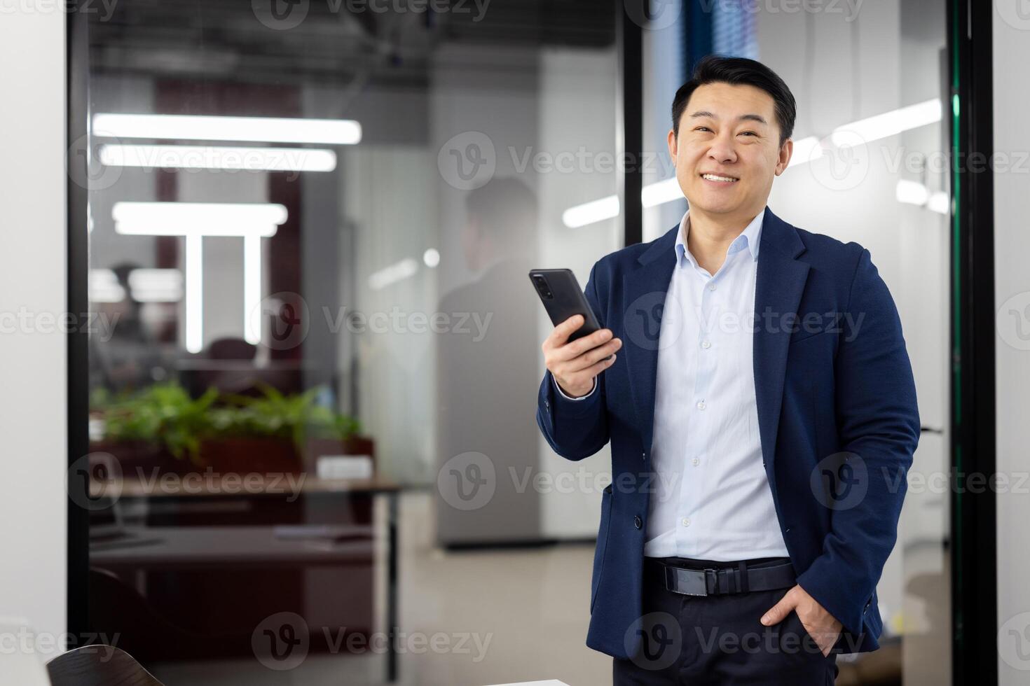 Portrait of successful young Asian man in suit standing in office, holding hand in pocket and mobile phone in other hand, looking confidently and smiling at camera. photo