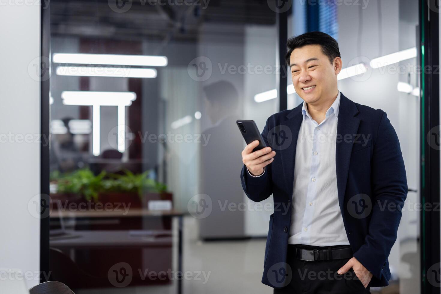 Asian businessman standing inside office with phone in hands, boss in business suit typing text message and reading social media, mature man using app on smartphone. photo