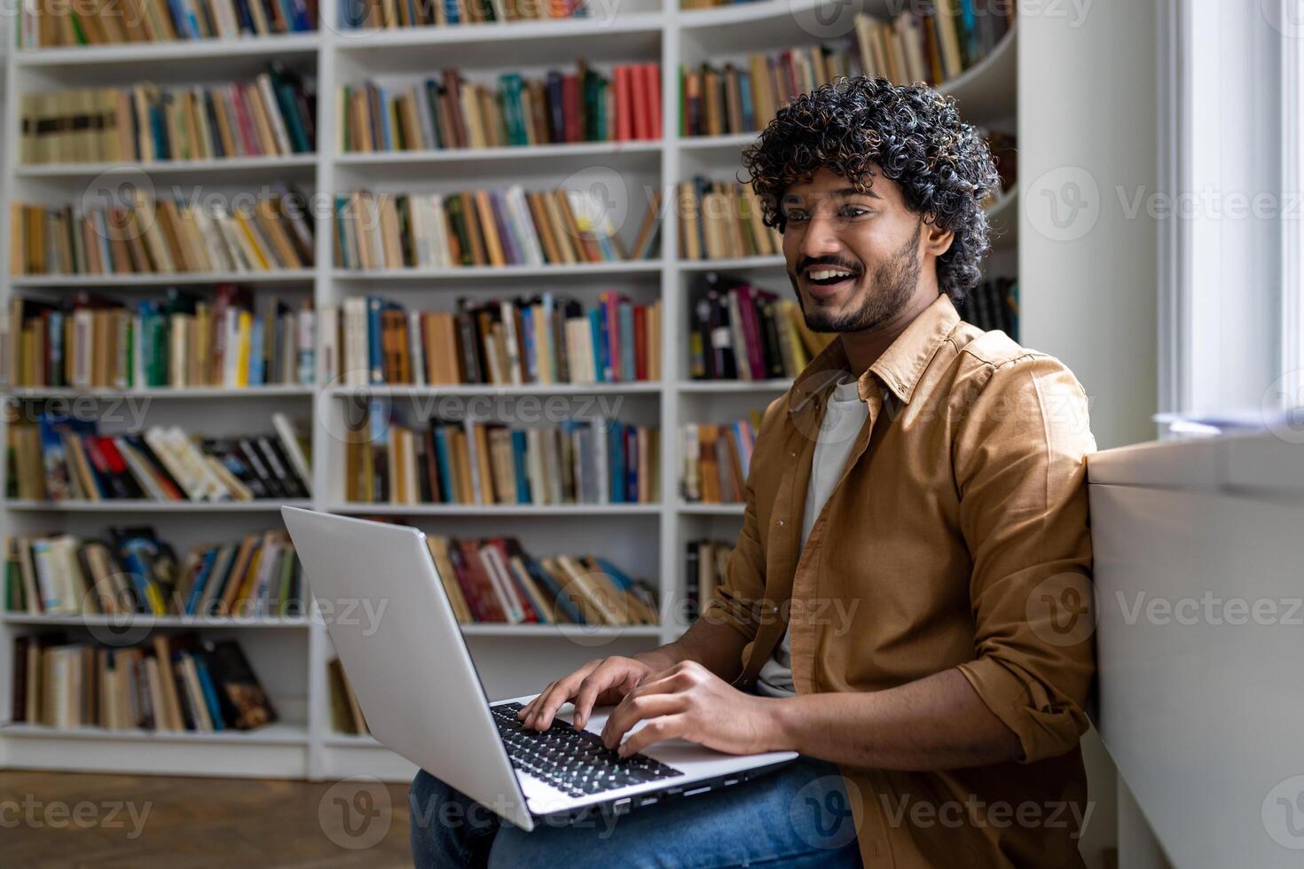Young hispanic student studying inside academic library among bookshelves, man typing on laptop keyboard, smiling contentedly sitting near window. photo
