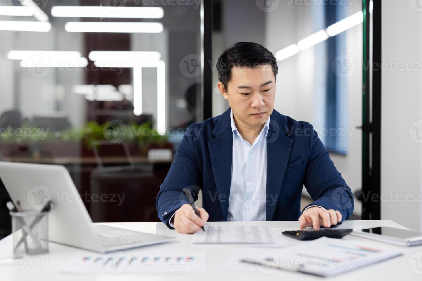 A professional businessman dressed in a blue suit is focused on reviewing financial documents at a modern workspace, surrounded by graphs and a laptop. photo