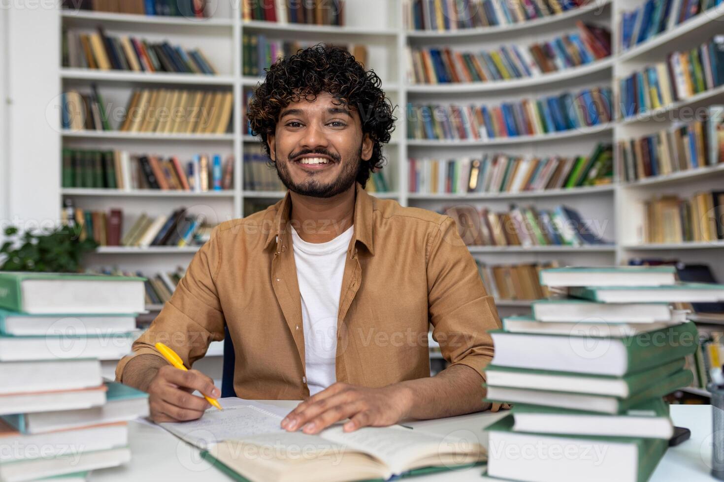 Happy and smiling student studying inside academic library, man smiling and looking at camera doing homework among books. photo