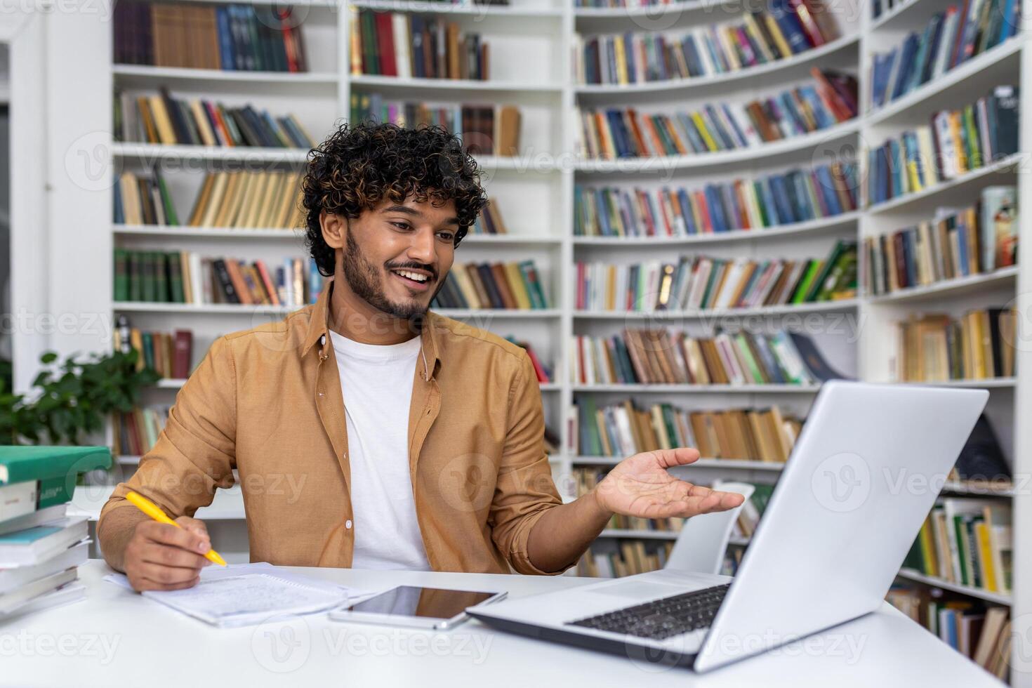 A young, curly-haired man smiles while gesturing at a laptop screen, working in a modern library setting. photo