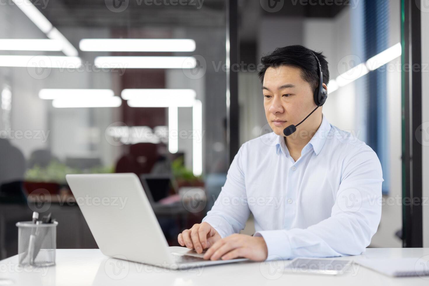 Focused Asian businessman with headset using laptop in a contemporary office setting, epitomizing professionalism and efficiency. photo