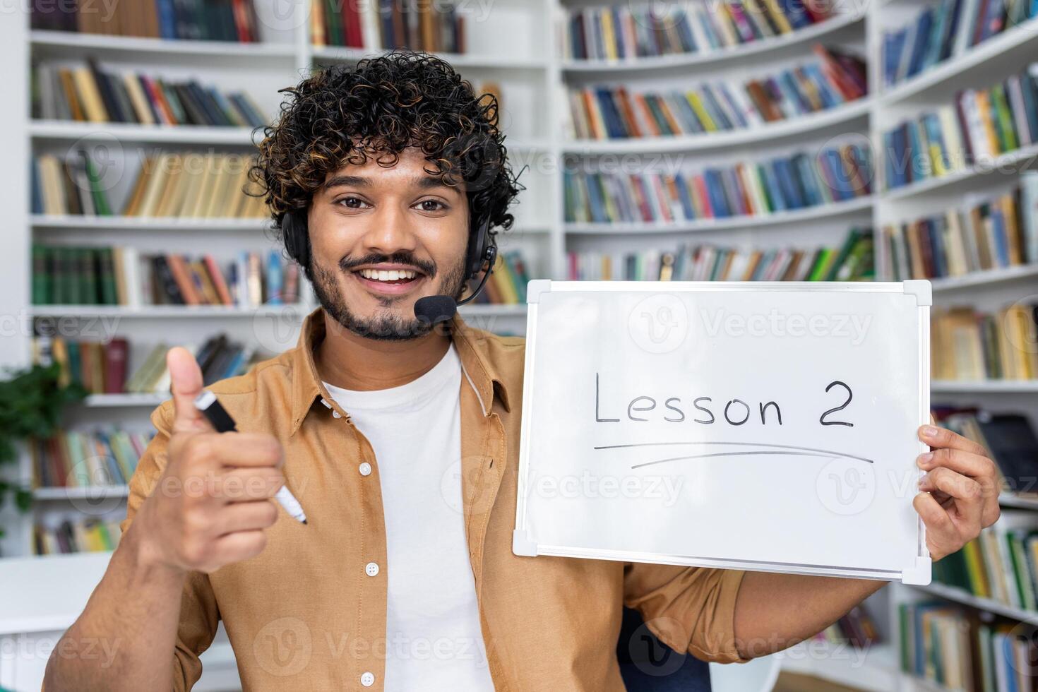Arab male holding whiteboard with inscription Lesson 2 on it and showing thumbs up gesture. Smiling online tutor wearing headphones and sitting on background of shelves with numerous of books. photo