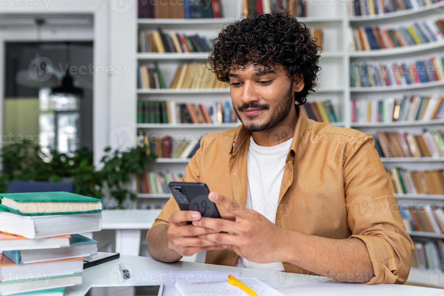 A cheerful young adult uses his smartphone amidst study materials, exuding a relaxed and happy vibe in a home environment. photo