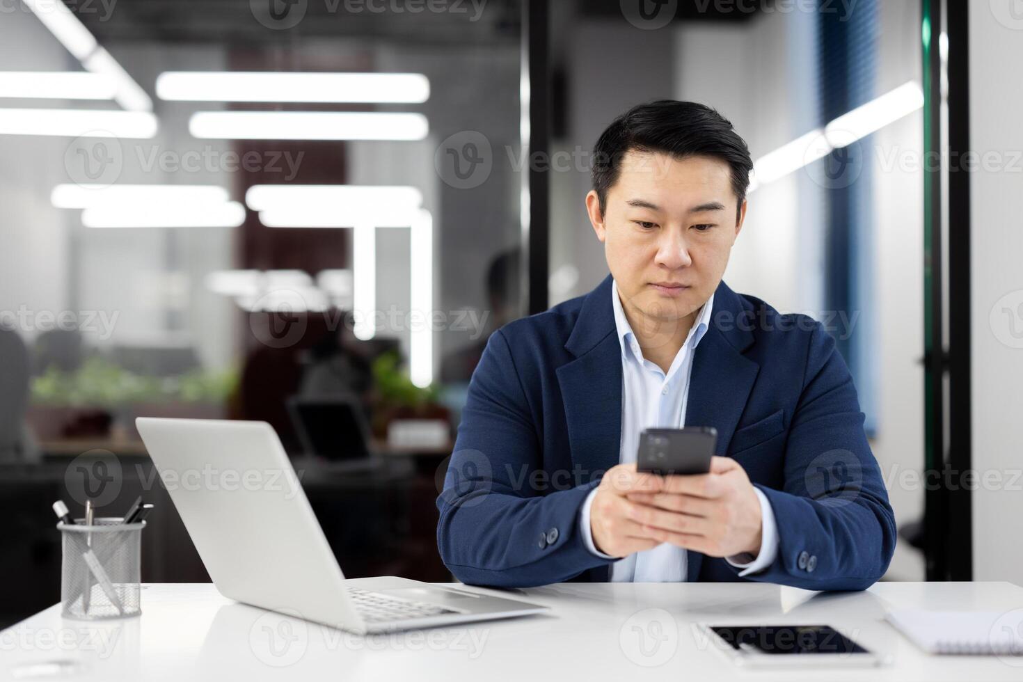 Serious Asian businessman sitting in the office at the desk with a laptop and using a mobile phone, texting, typing messages. photo