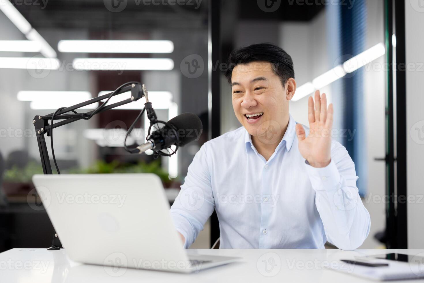 Asian smiling young man sitting in office at desk in front of microphone and laptop and having online meeting via call, waving hand and greeting to camera. photo