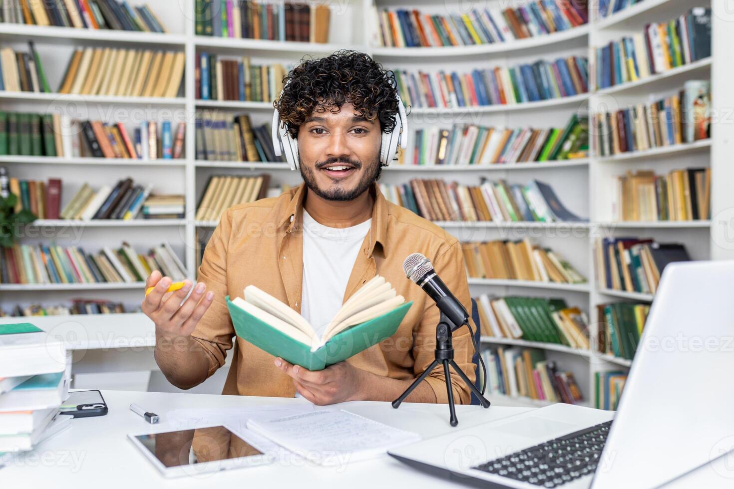 Confident blogger in white headset recording audio content while sitting with daybook by desk with professional micro. Islamic lecturer reading information for remote listeners in cyberspace. photo