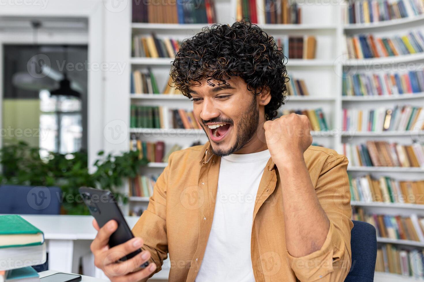 Shocked indian man working at desk surrounded by books shelves and technology. Surprised male freelancer holding smartphone in hand and appearing to reading positive news indoors. photo