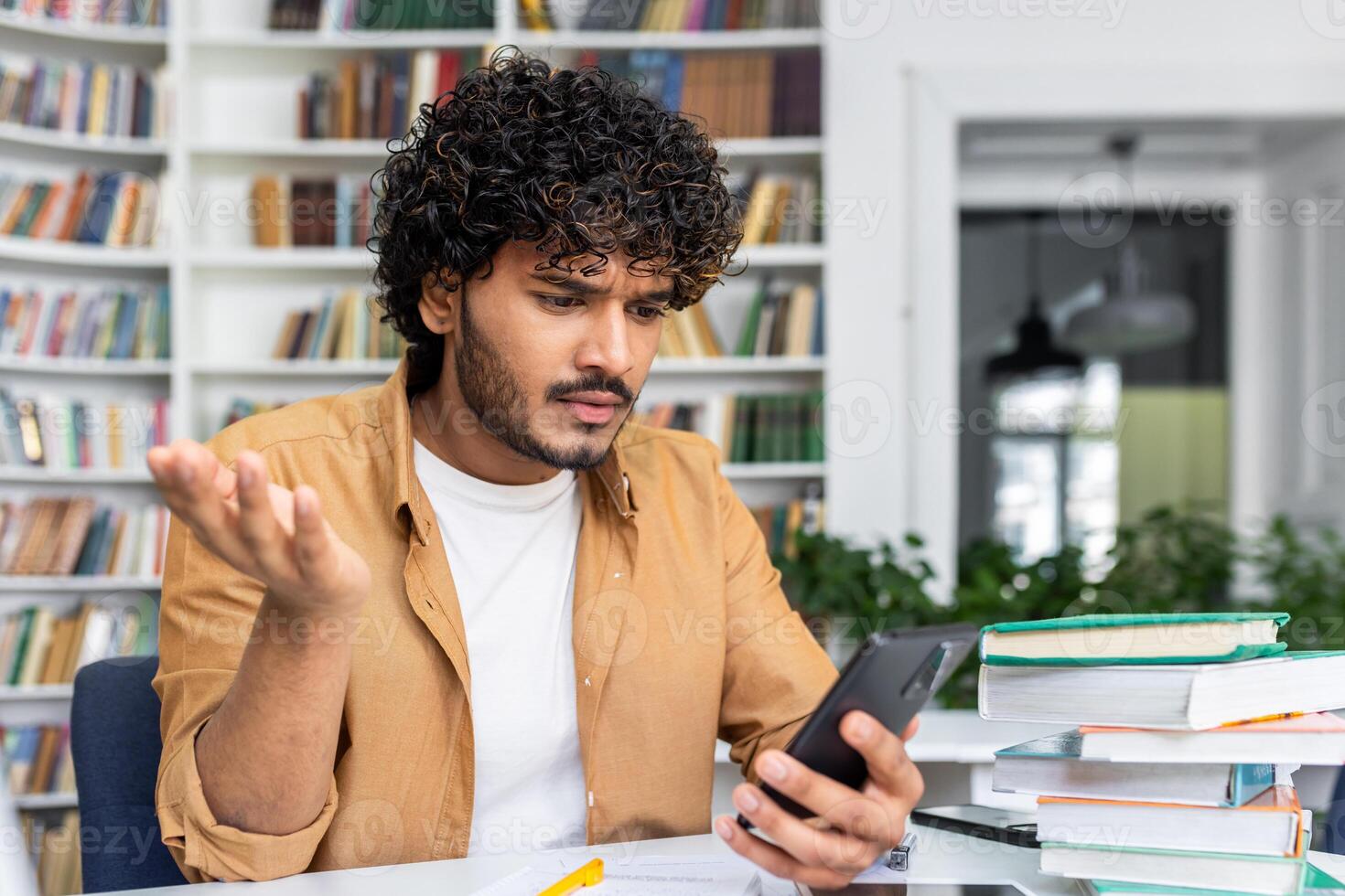 A young adult man looks puzzled while reading something on his smartphone, surrounded by a stack of books, possibly studying. photo