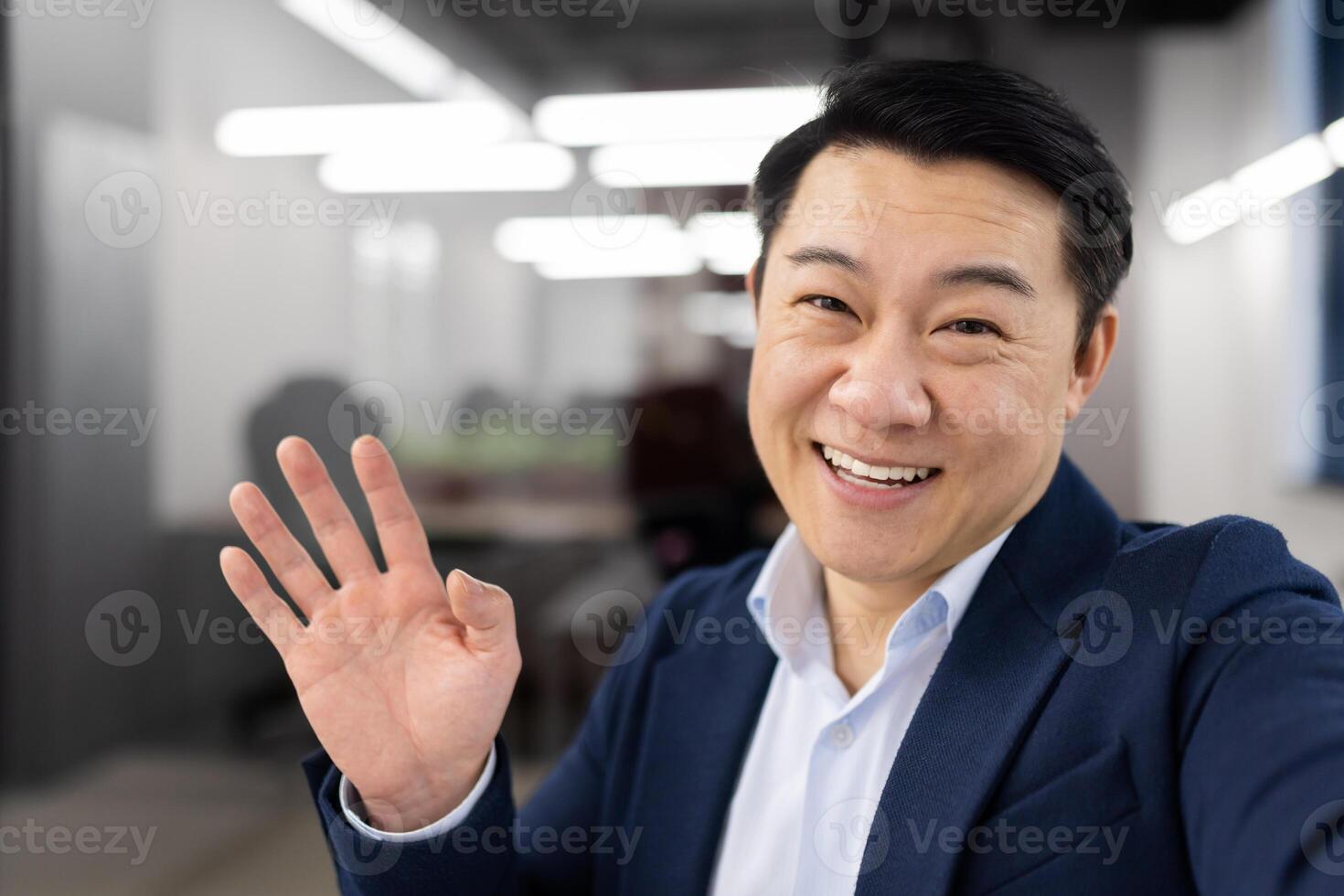 Friendly japanese employer in strict outfit waving hello while looking at camera with toothy smile. Positive entrepreneur connecting to group meeting by digital gadget with newly hired trainees. photo