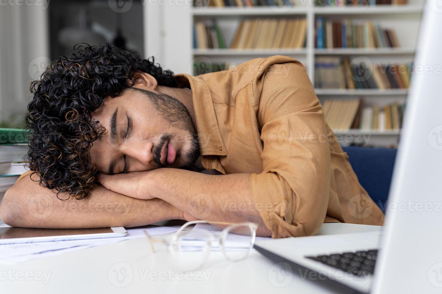 Tired student sleeping on table inside university academic library, hispanic man among books with laptop preparing for session and final exams. photo