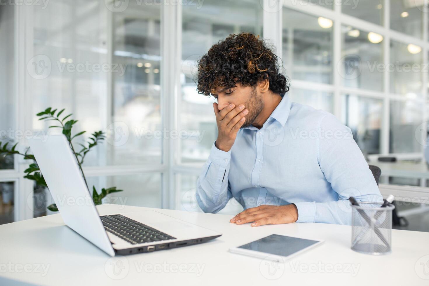 Young man feeling nausea while working at his desk in a modern office, indicating discomfort or illness, possibly related to stress or fatigue. photo