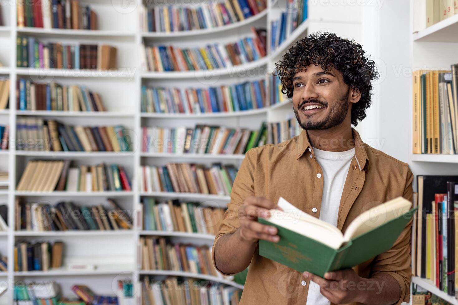 Young hispanic student reading a book while standing inside an academic university library, man smiling contentedly looking towards the window. photo