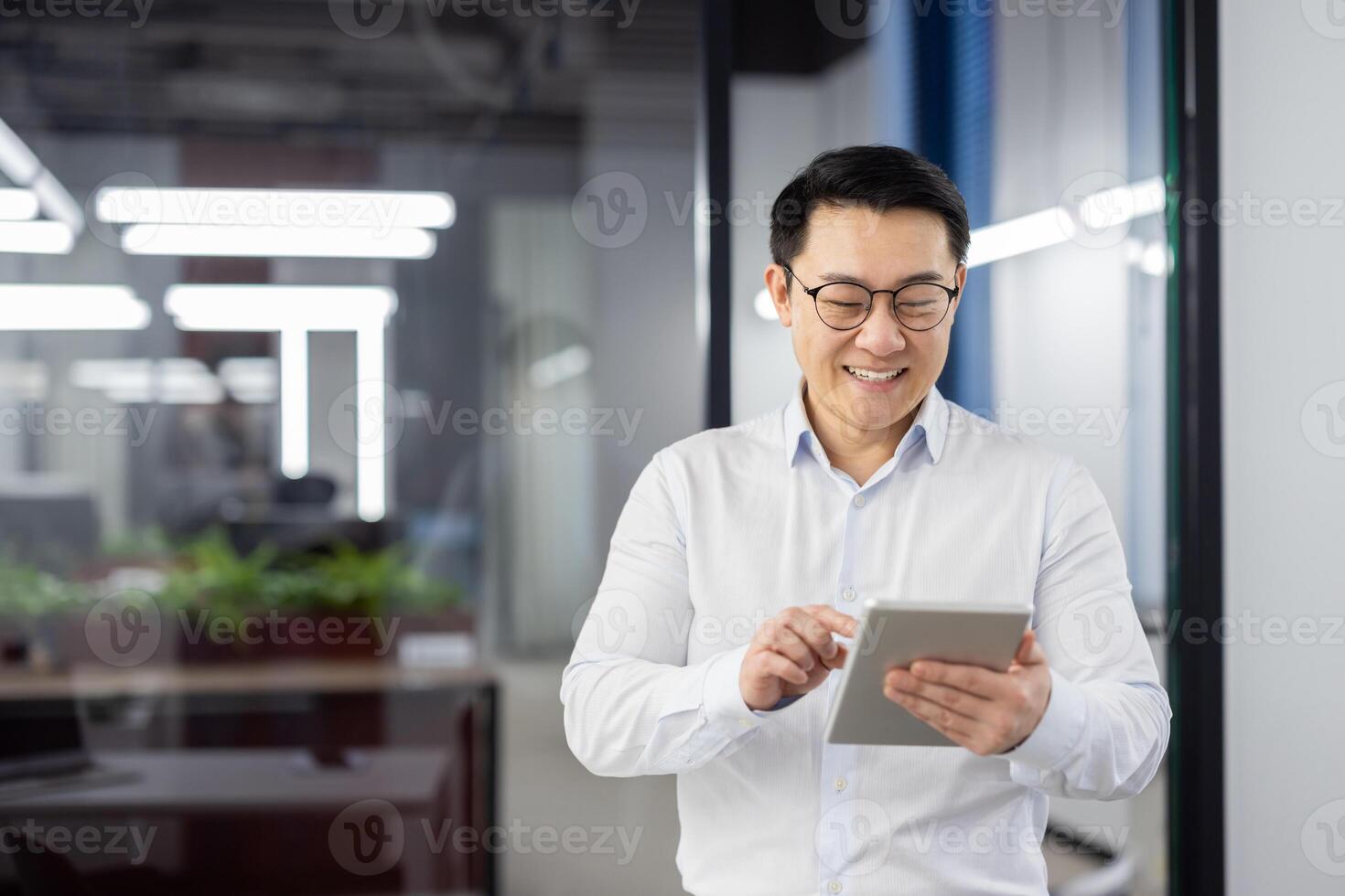 Asian young man working in office, standing inside and using tablet, looking at screen smiling and typing. photo