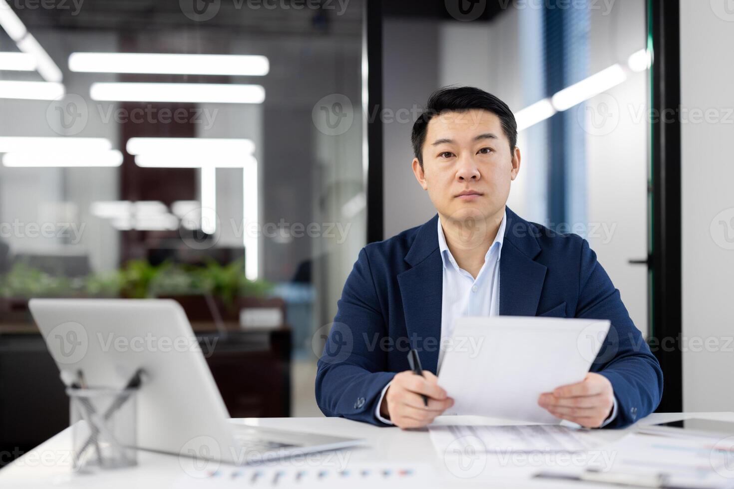 Focused Asian businessman sitting at desk in a well-lit office, reviewing documents with serious expression, displaying professionalism and determination. photo