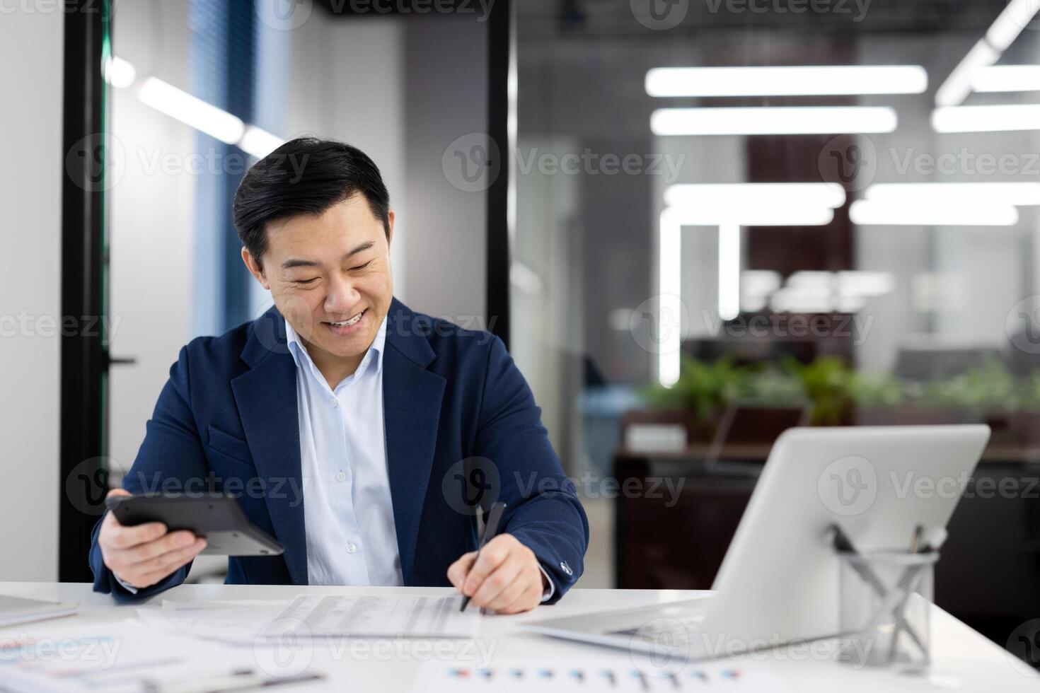 A professional Asian businessman reviews financial documents while smiling in a brightly lit modern office environment, portraying efficiency and contentment. photo