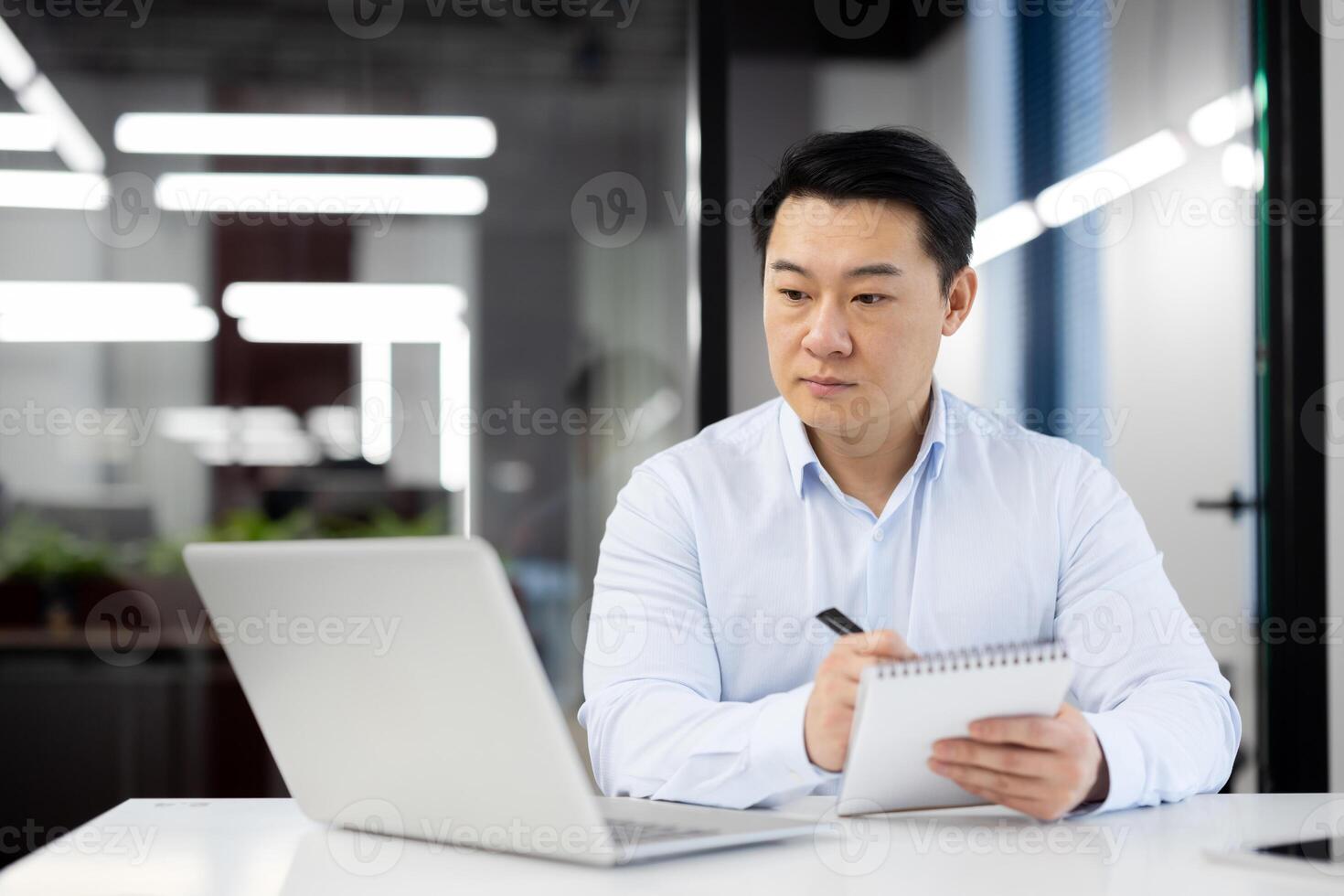 Asian business man watching training course online, man inside office at workplace writing in notebook, using laptop for remote communication with mentor and coach. photo