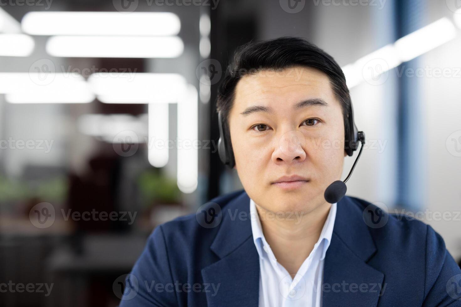 Close-up portrait of a serious young Asian man wearing a headset sitting in the office and looking confidently into the camera. photo