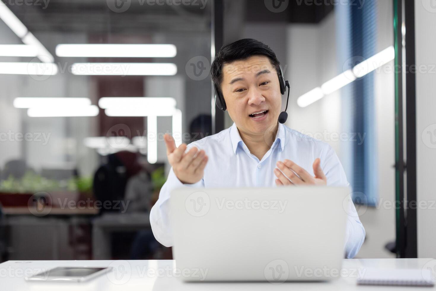 Cheerful Asian businessman conversing on a call with a laptop and headset in an office setting. photo