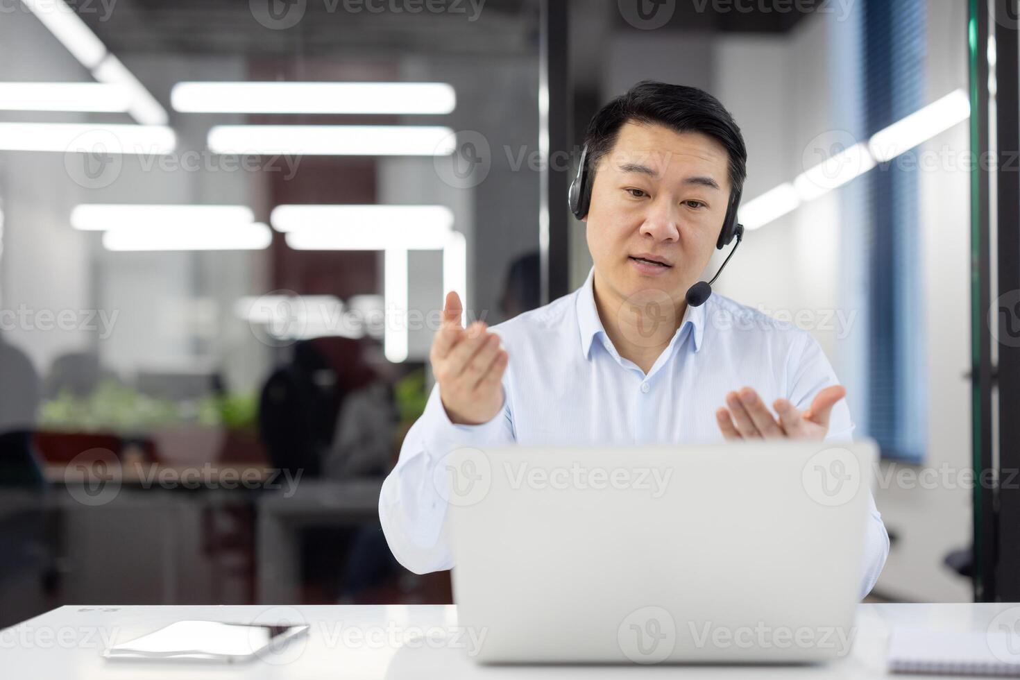 Asian young man in a white shirt and a headset sits in the office at the desk in front of the laptop and talks, conducts a consultation via call, gesturing with his hands. photo
