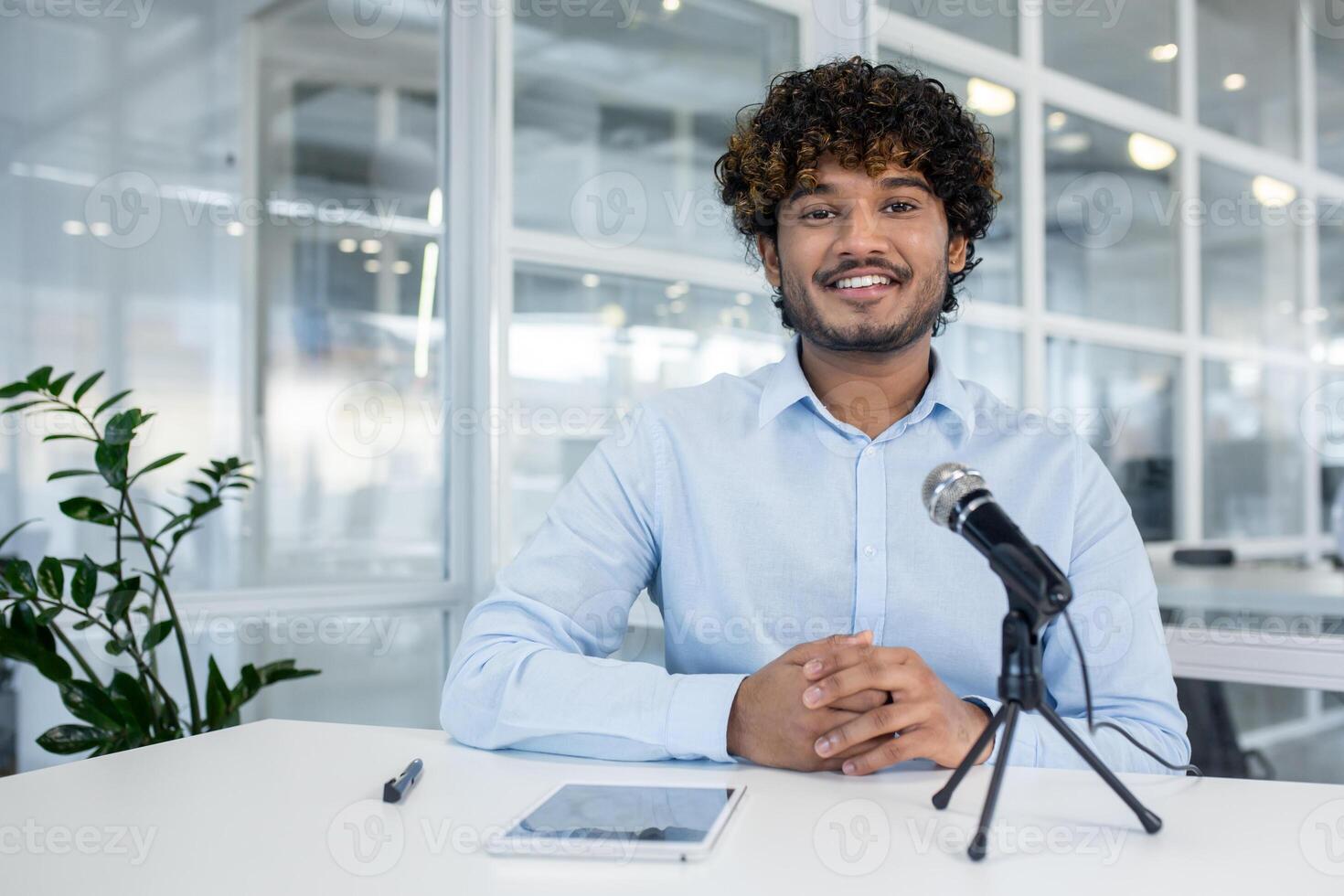 Smiling young man with curly hair dressed in a light blue shirt engaging in a call using a microphone ,and tablet in a modern office. photo