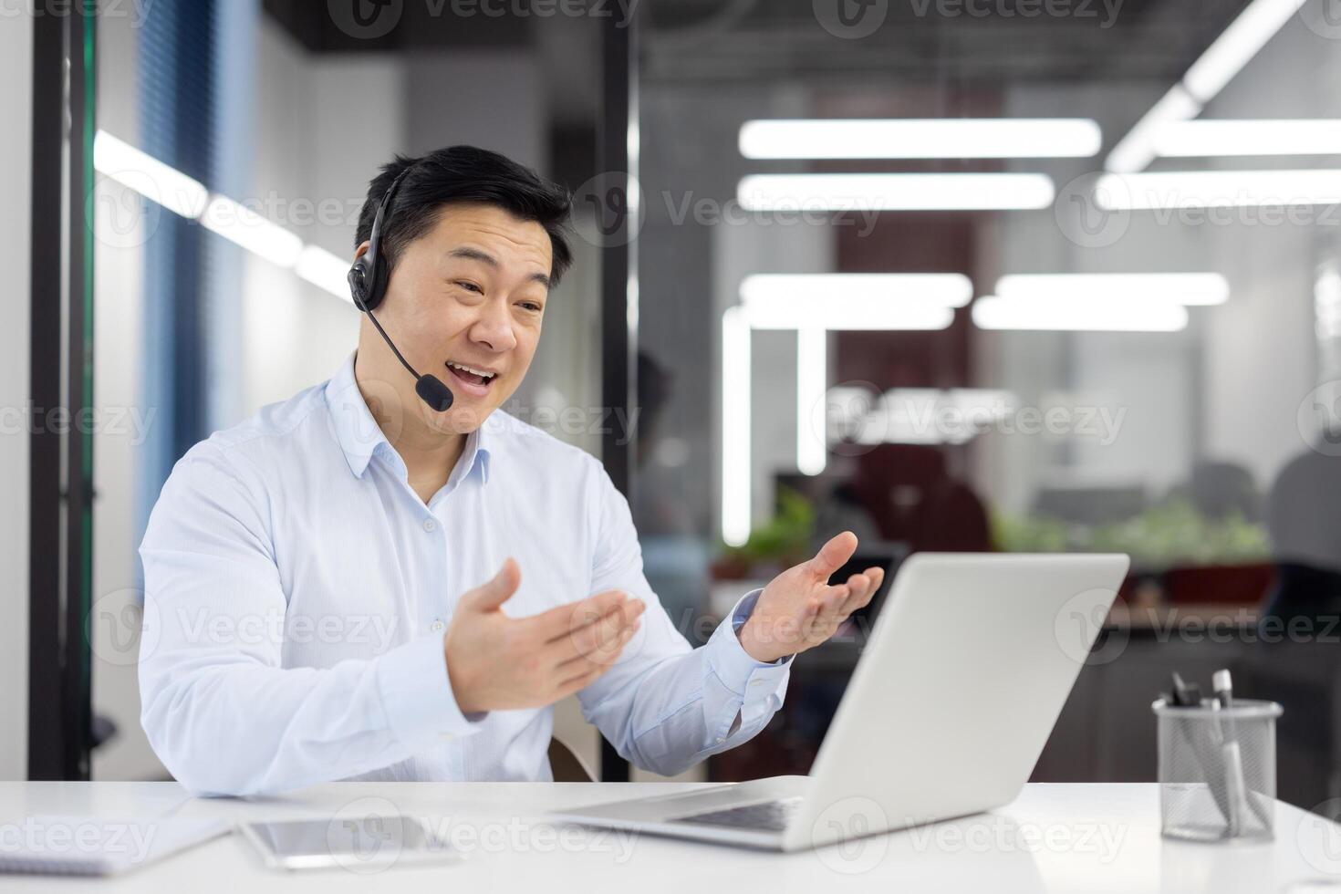 Smiling asian man in white shirt working in office at desk, wearing headset, talking on call through laptop gesturing with hands. photo