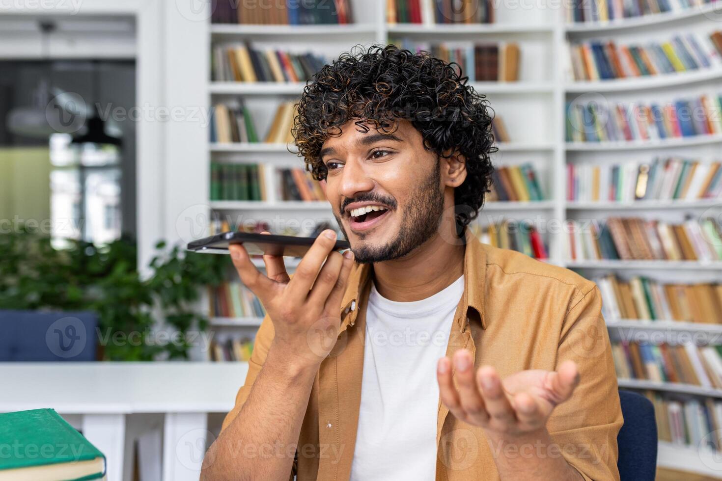 Cheerful young man with curly hair speaks into his smartphone using voice command in a library setting. photo