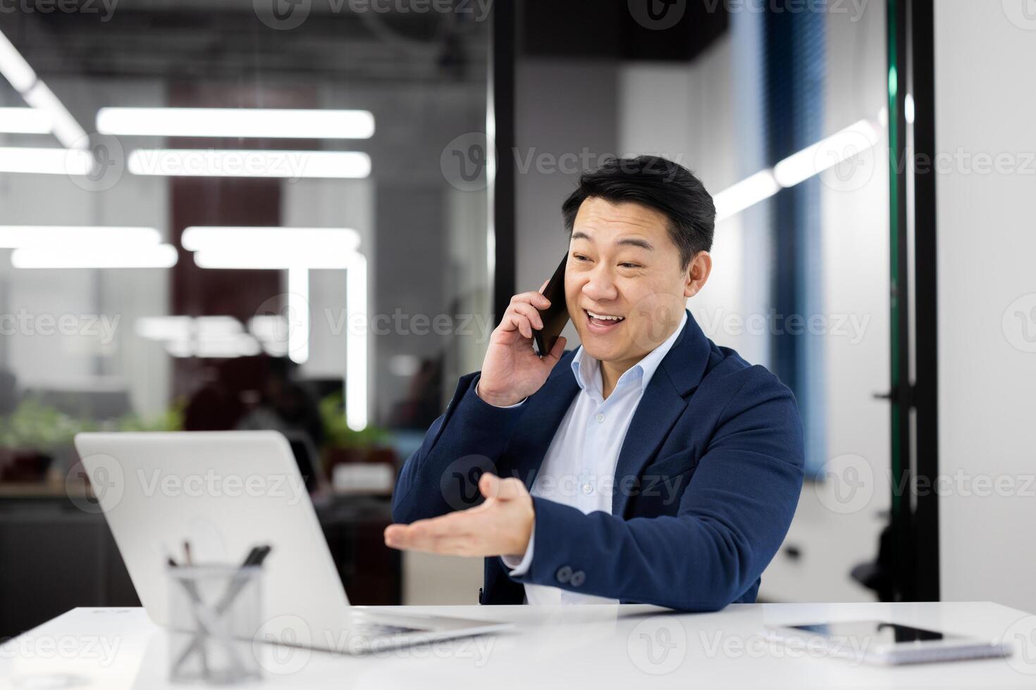 Millennial male sitting alone at workspace and talking by cellphone while spreading hand to portable computer. Friendly manager having remote conversation with colleague about project development. photo