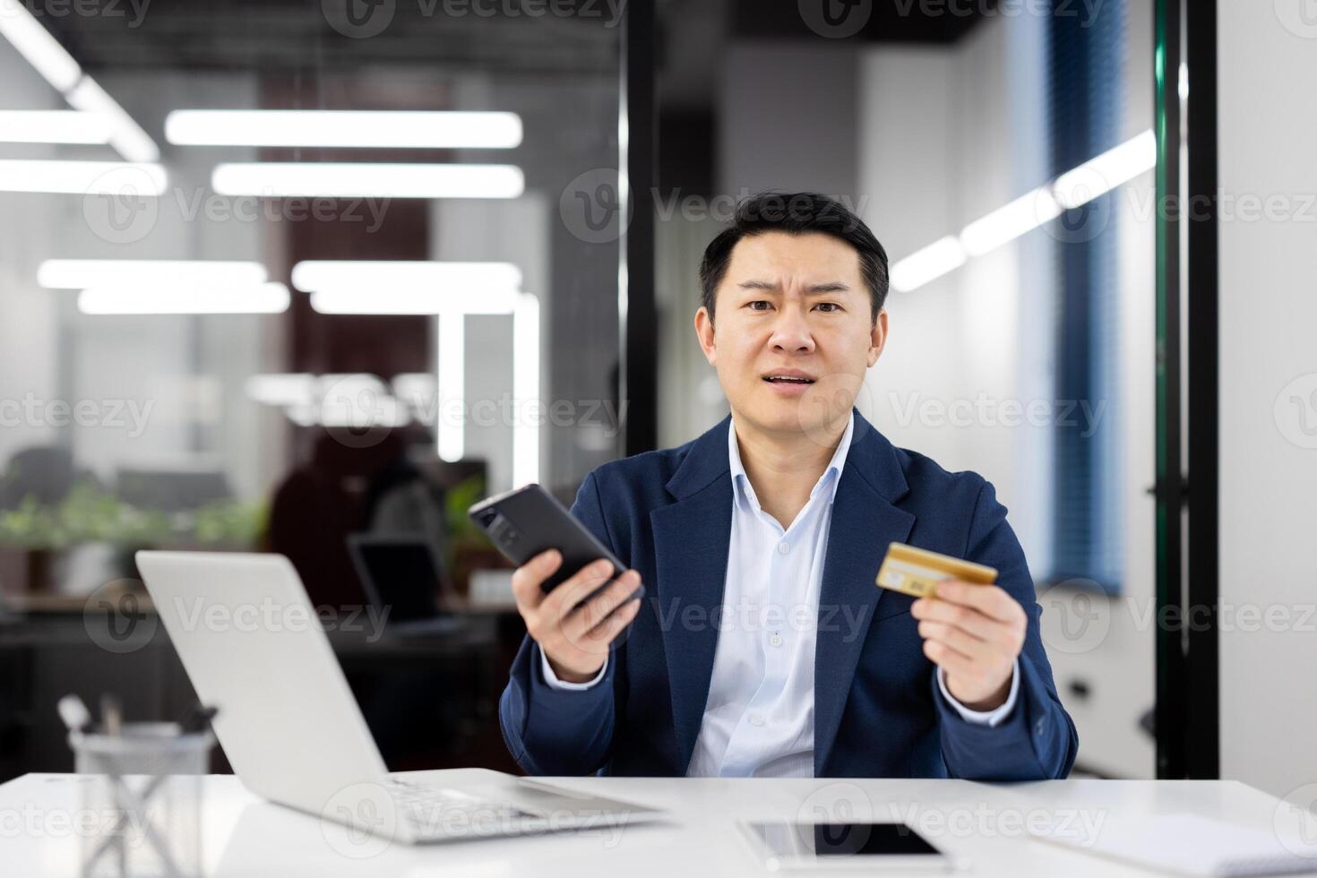 A businessman in a modern office looks perplexed, holding a smartphone and a credit card. Concept of financial security and potential fraud concerns. photo