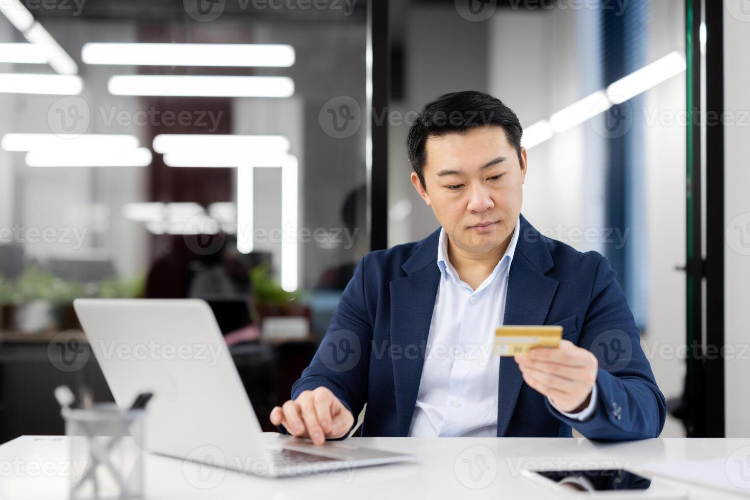 Focused Asian businessman in a modern office making a payment online using a credit card. This professional scene emphasizes e-commerce and secure transactions in a corporate setting. photo
