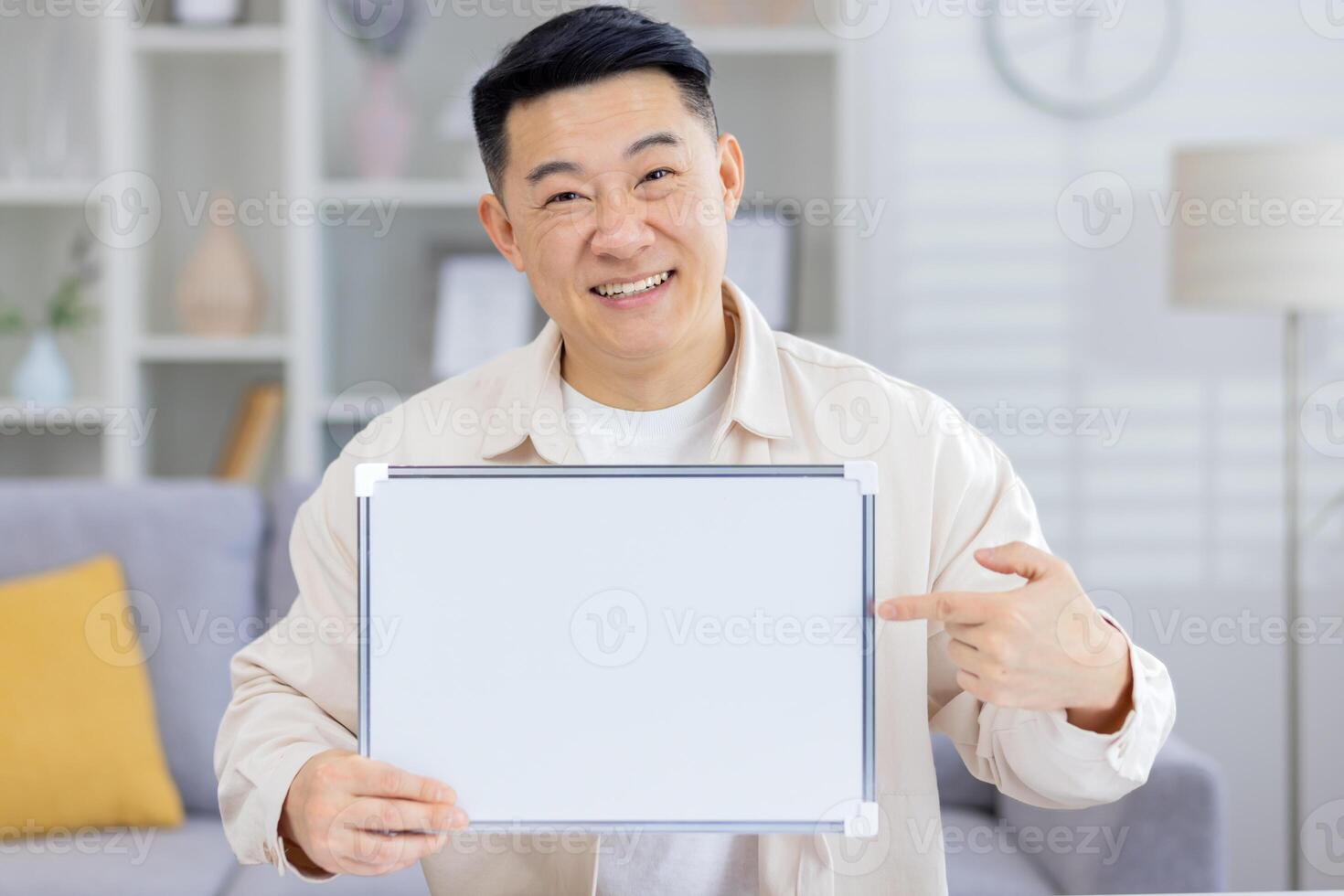 Smiling man holding a blank sign while pointing and looking cheerful in a cozy living room. Perfect for messages and advertising concepts. photo