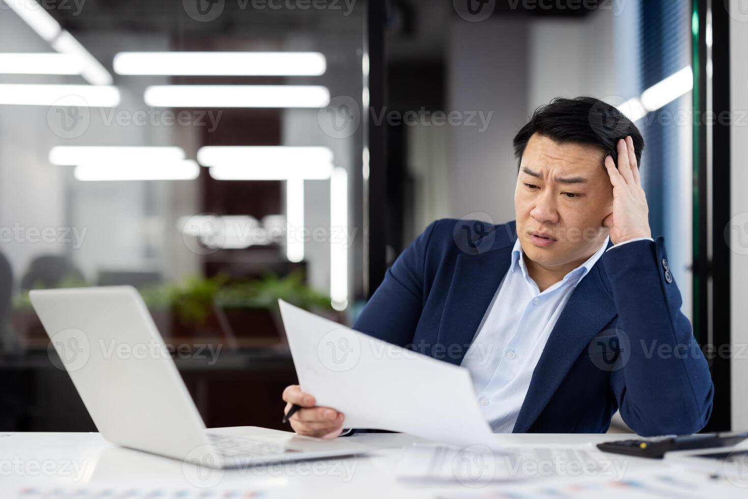 Confused korean male working with documents in front of portable laptop in office environment. Exhausted businessman dressed in dark suit reading important papers indoors. photo