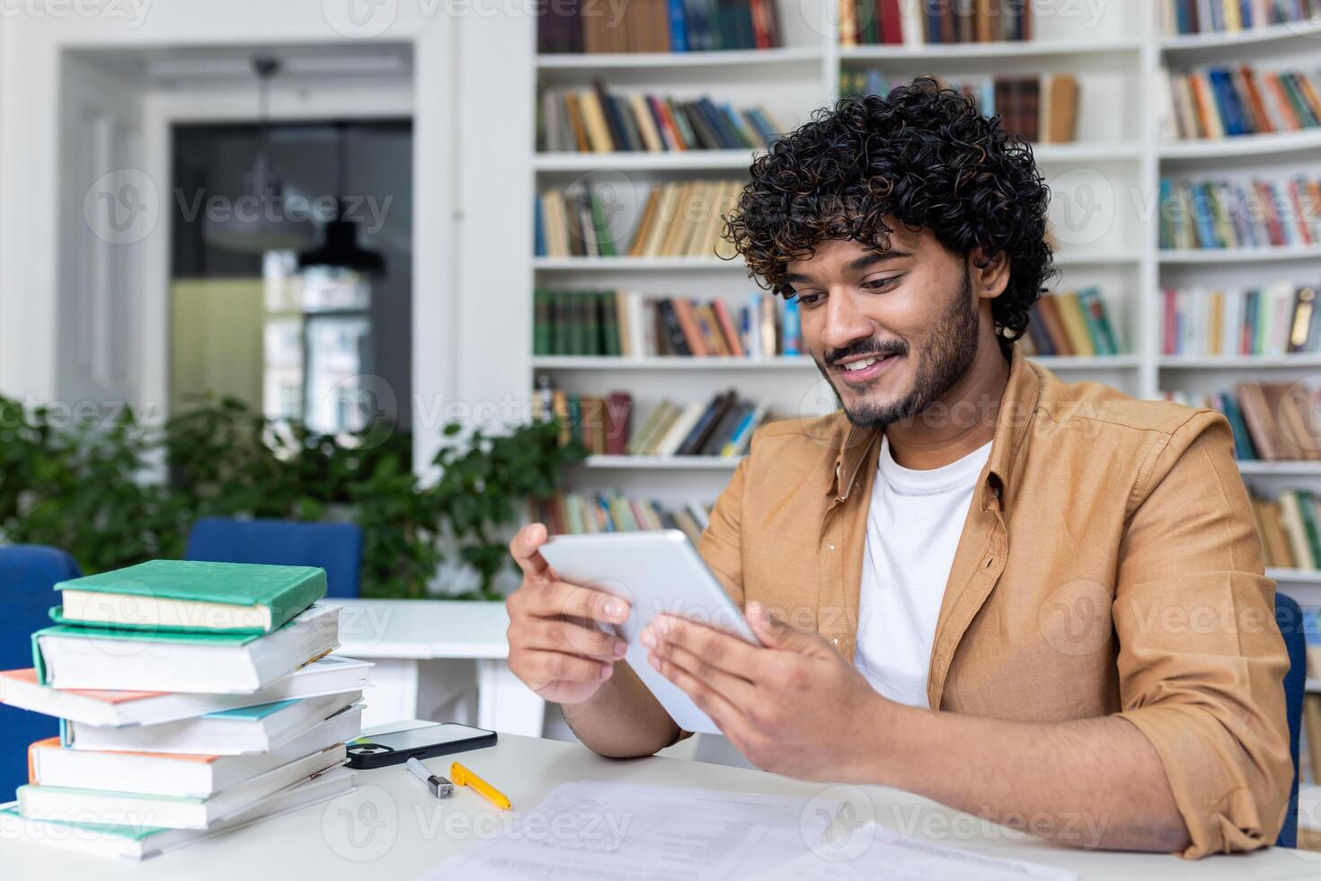 Young successful student studying sitting inside university academic library, man smiling holding tablet computer, Hispanic man using study app. photo