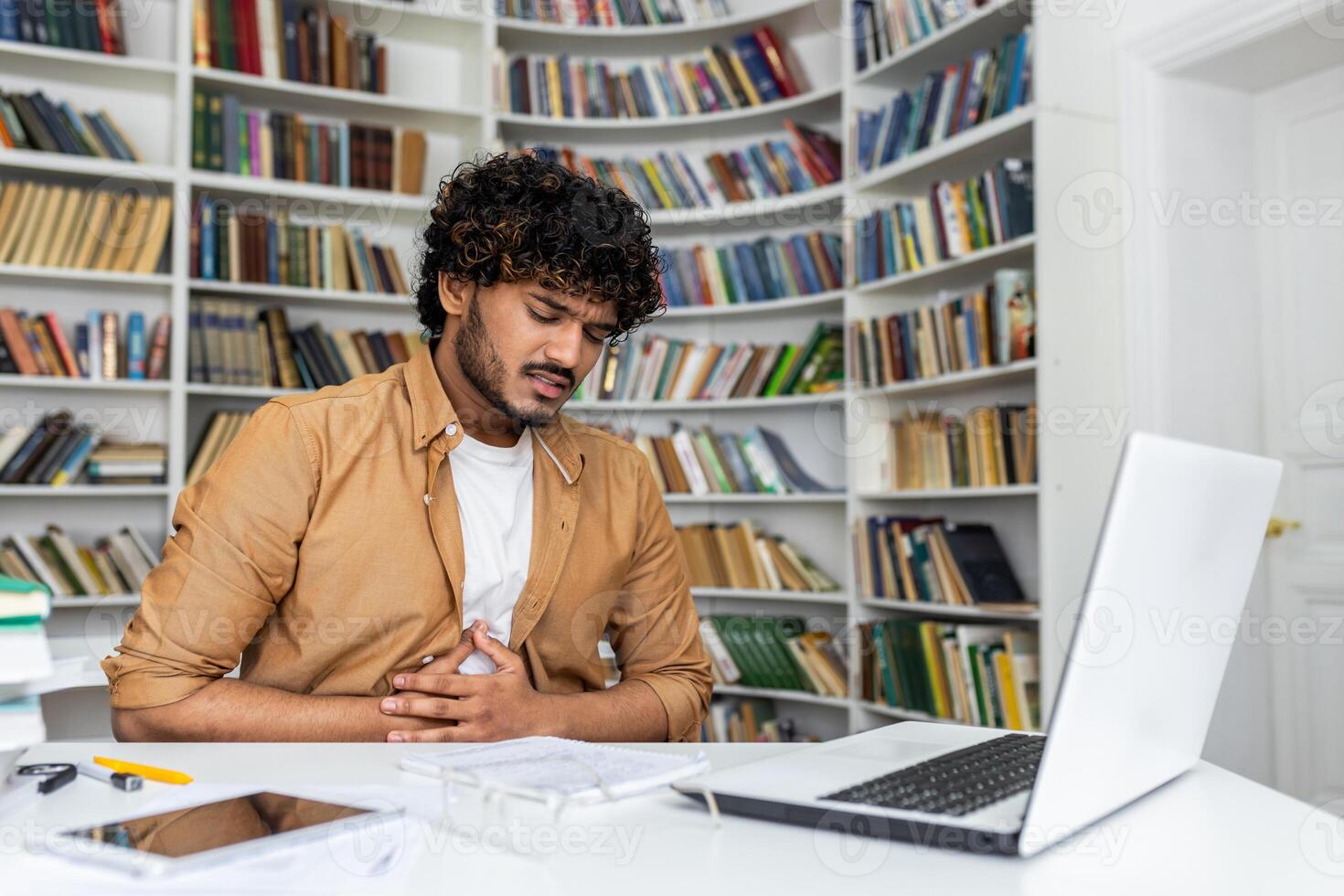 Unhealthy diverse guy looking down and keeping arms on belly while spending day at public library with pc. University student struggling with digestion ache and inability of concentrating on studying. photo